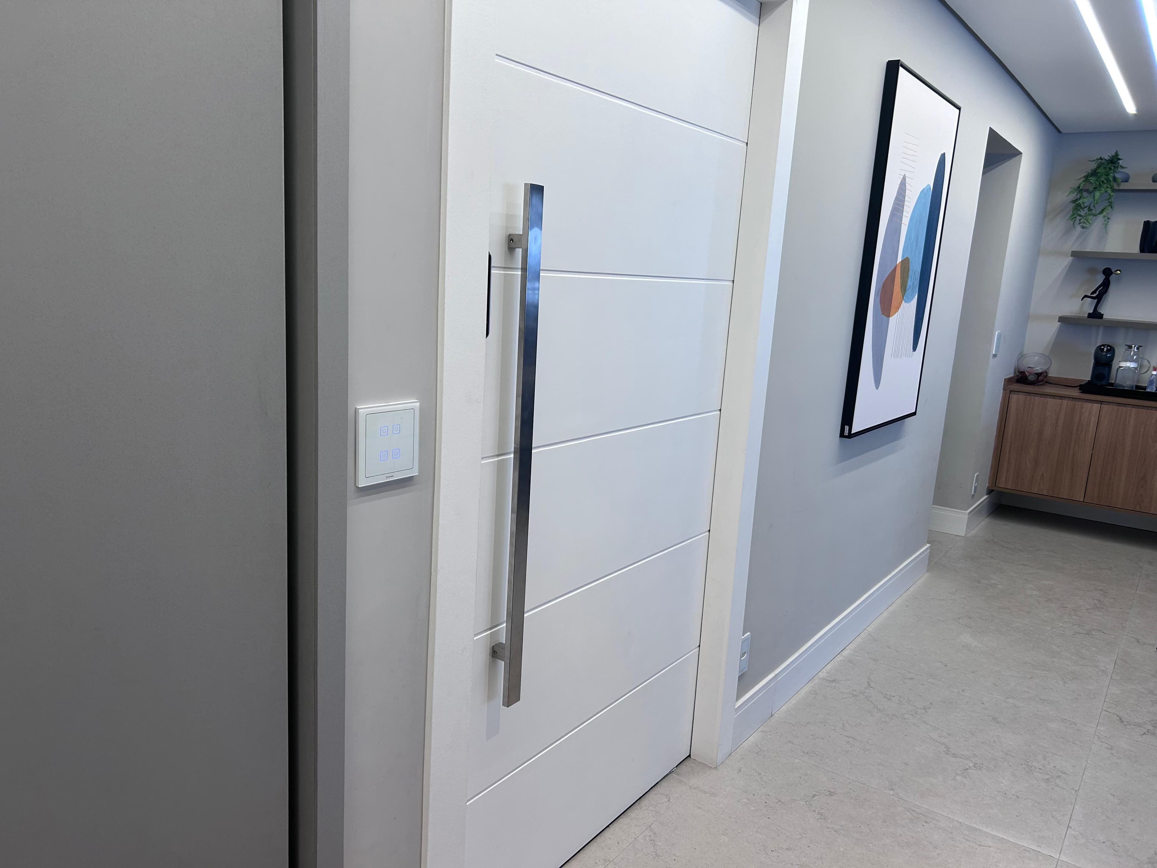 Corredor de residência com um interruptor Outmat de quatro teclas na ombreira da porta, luminária linear no teto e vista até o fundo do ambiente.